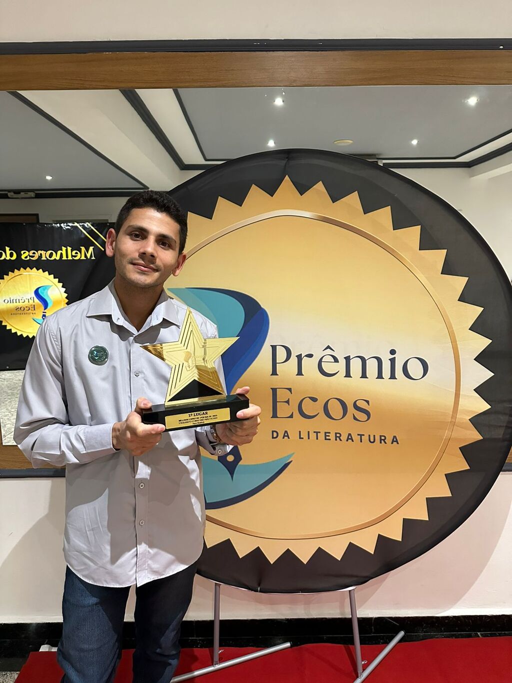 homem de camisa social e calça jeans exibe trofeu em frente a painel com inscrição 7ª Edição do Prêmio Ecos de Literatura