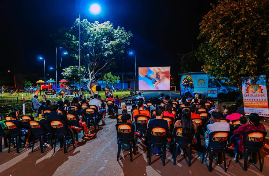 plateia assiste desenho em tela grande montada numa praça, ao lado de um furgão