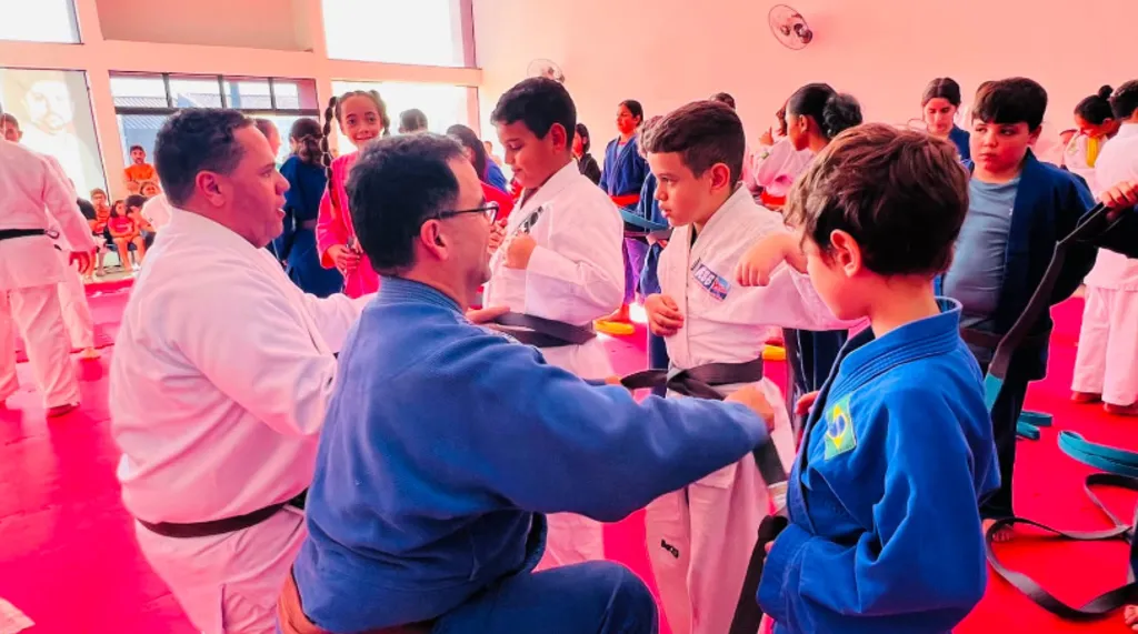 Instrutores em um tatame vermelho ajustando as faixas no kimono das crianças durante um treino ou graduação de judô.