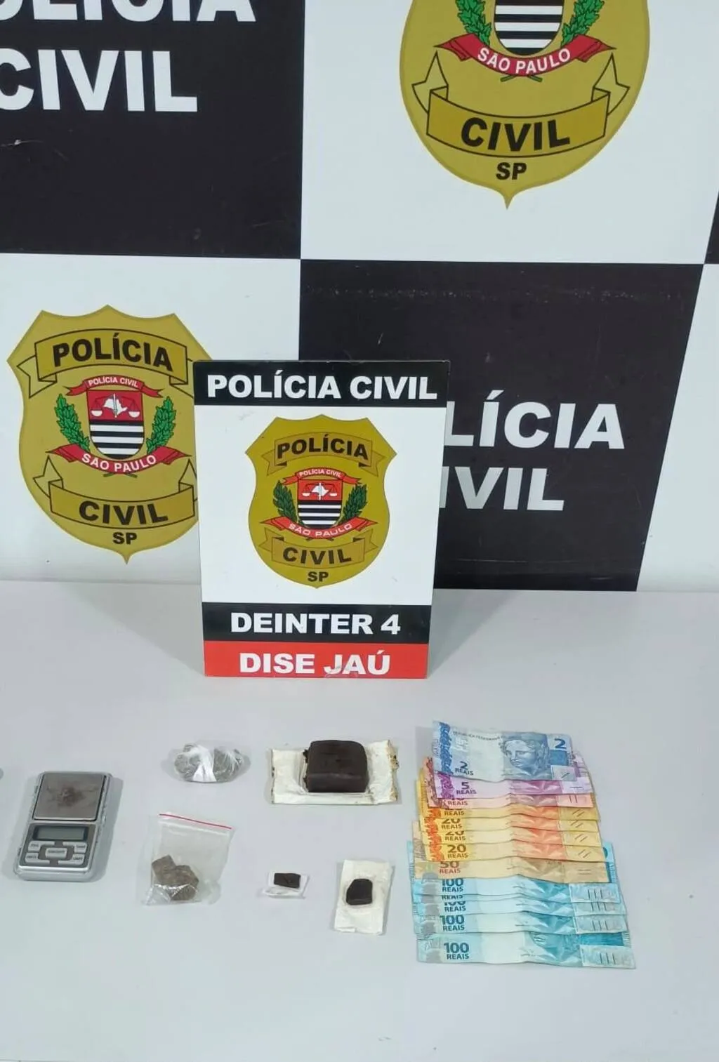 Mesa contendo dinheiro, balança e porções de entorpecentes à frente de um painel da Polícia Civil