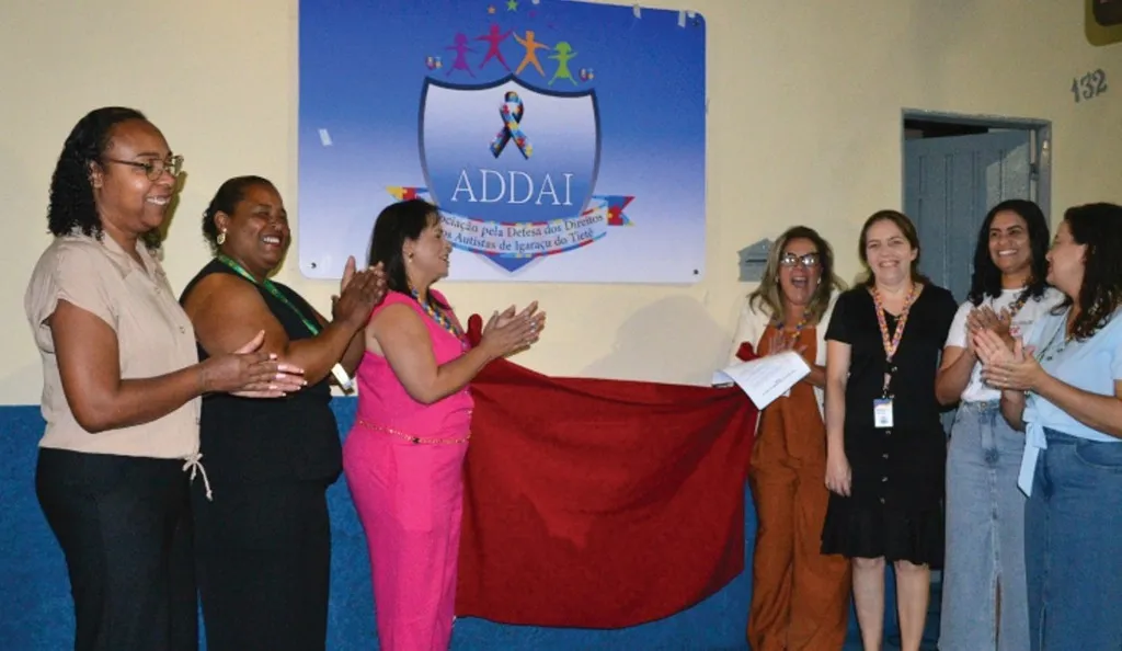Mulheres seguram um pano vermelho tendo ao fundo placa com símbolo autista escrita ADDAI