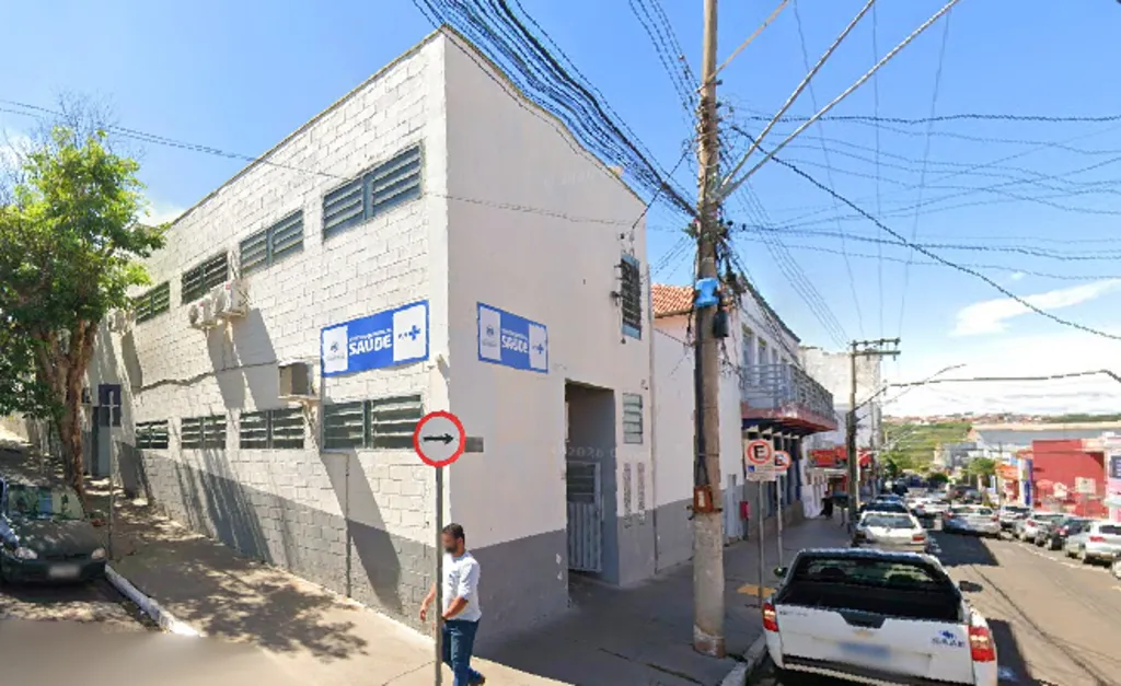 Prédio com placa escrito "Saúde", postes de energia elétrica e veículos estacionados nas laterais da rua