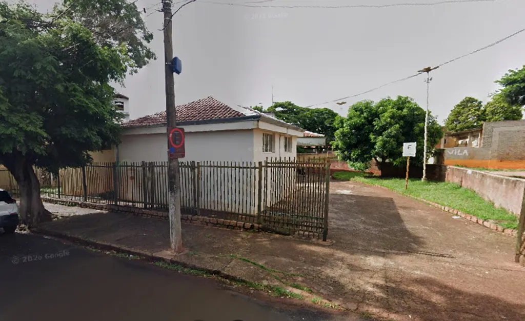 Prédio com janelas na lateral e paredes na cor branca, tendo a frente grade e um poste de energia elétrica na calçada