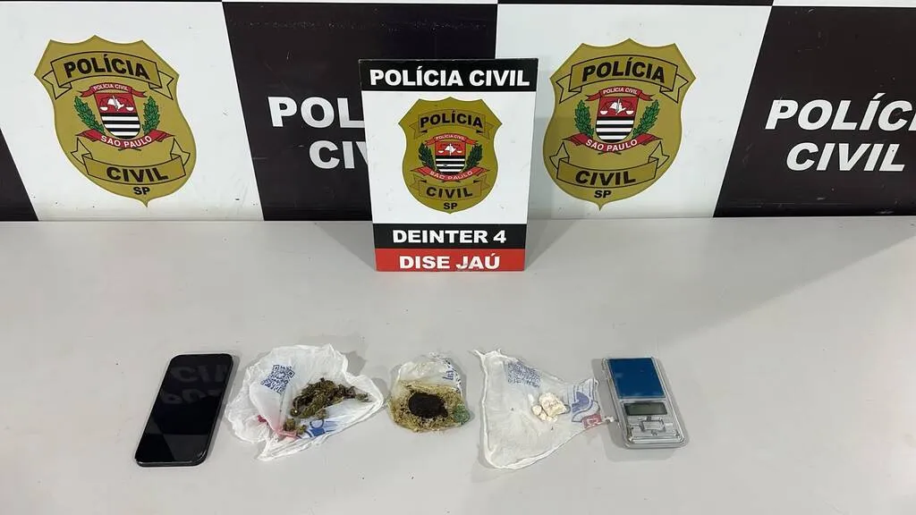 Telefone celular, drogas e balança colocados em uma mesa com painel da Polícia Civil ao fundo