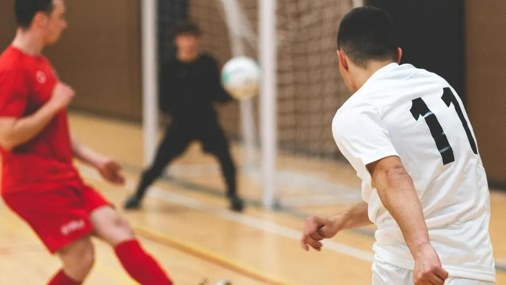 jogadores observam bola a caminho do gol em quadra de futsal