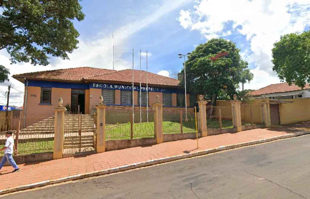 Fachada de escola com grades e gramado na fachada, tendo um homem de calça azul e camisa branca caminhando na calçada