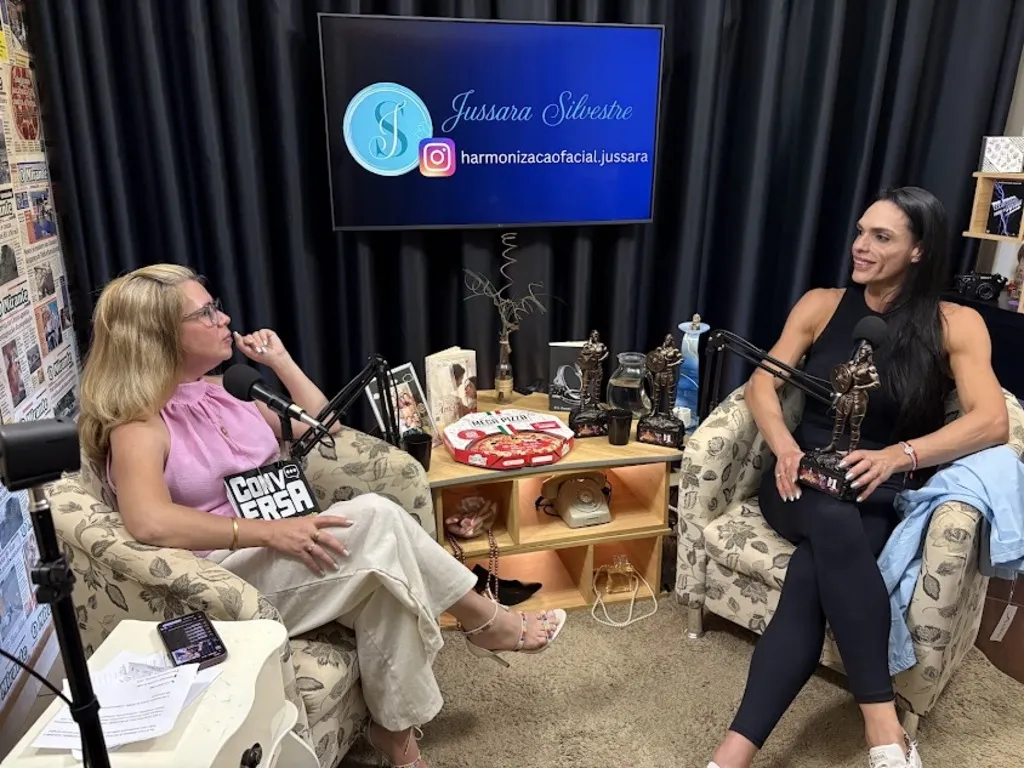 Duas mulheres estão gravando um podcast em um estúdio aconchegante, sentadas em poltronas floridas e com microfones à frente. A apresentadora de rosa à esquerda escuta atentamente a convidada de preto à direita, que sorri enquanto segura um pequeno troféu.