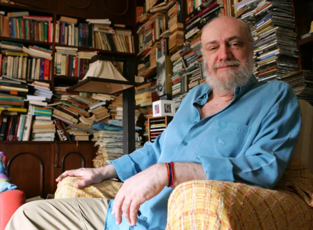 homem de barba branca e camisa azul sentado numa poltrona, em frente a uma estante de livros