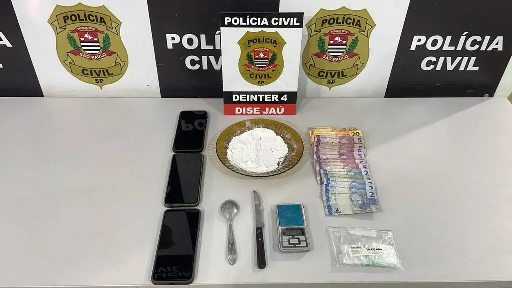Mesa com três telefones celulares, prato contendo pó branco, colher, faca, balança e dinheiro, além de painel da Polícia Civil ao fundo