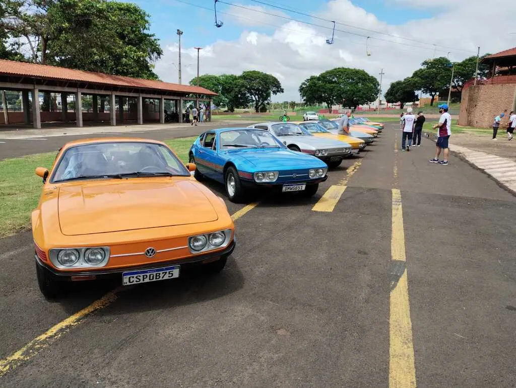 Carros modelo VW SP2 de cores variadas perfilados do lado esquerdo, com pessoas a pé e asfalto com faixas amarelas