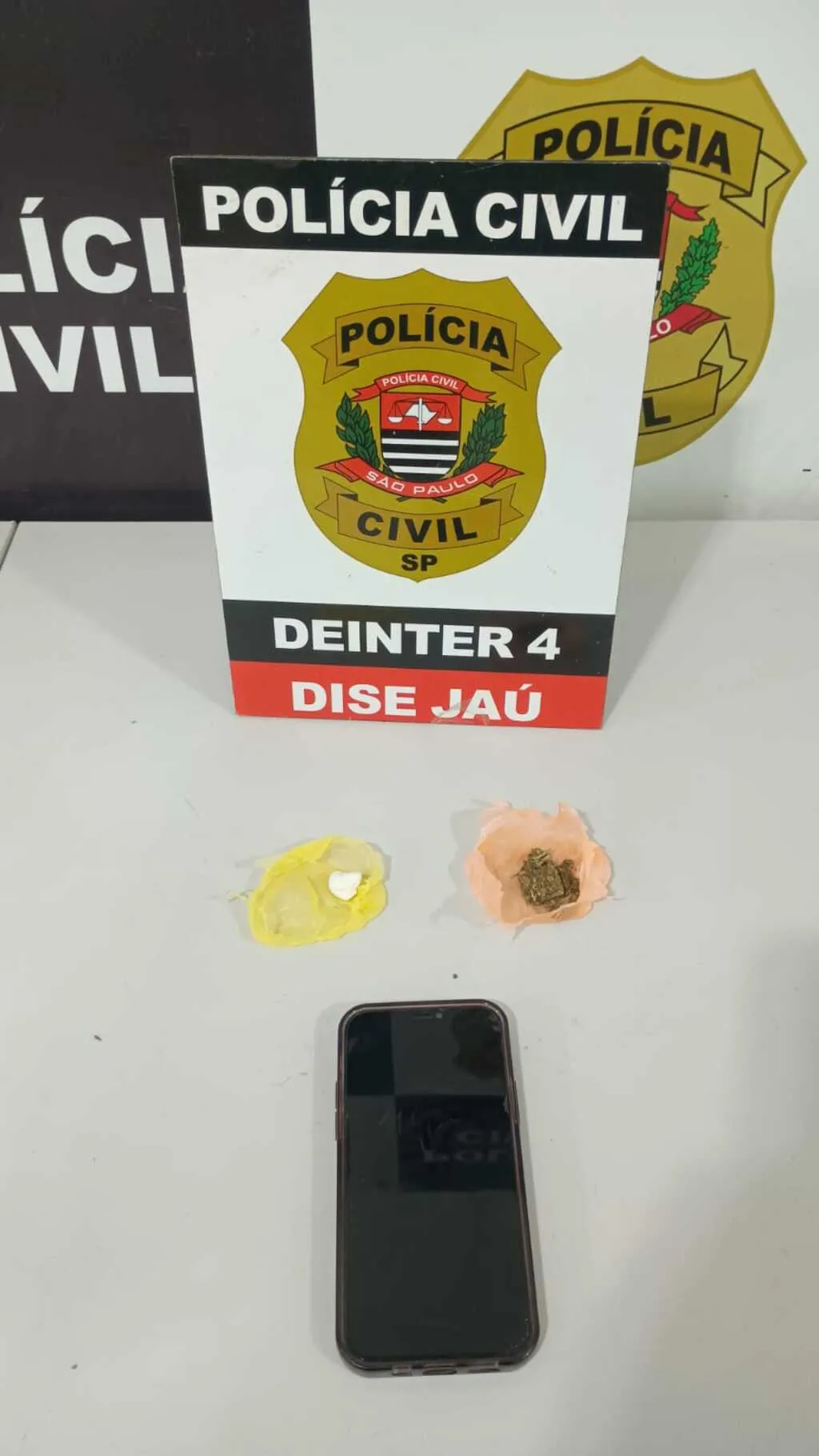 Celular, porções de drogas e um painel com o símbolo da Polícia Civil do Estado de São Paulo ao fundo