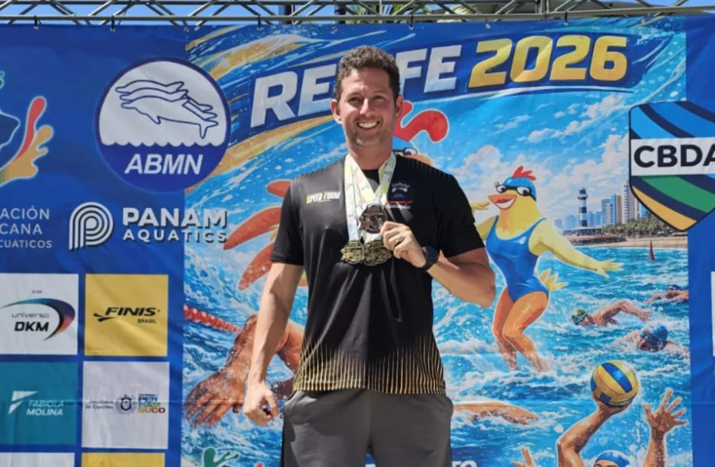 homem exibe medalhas em frente a um painel com patrocinadores e elementos de píscina