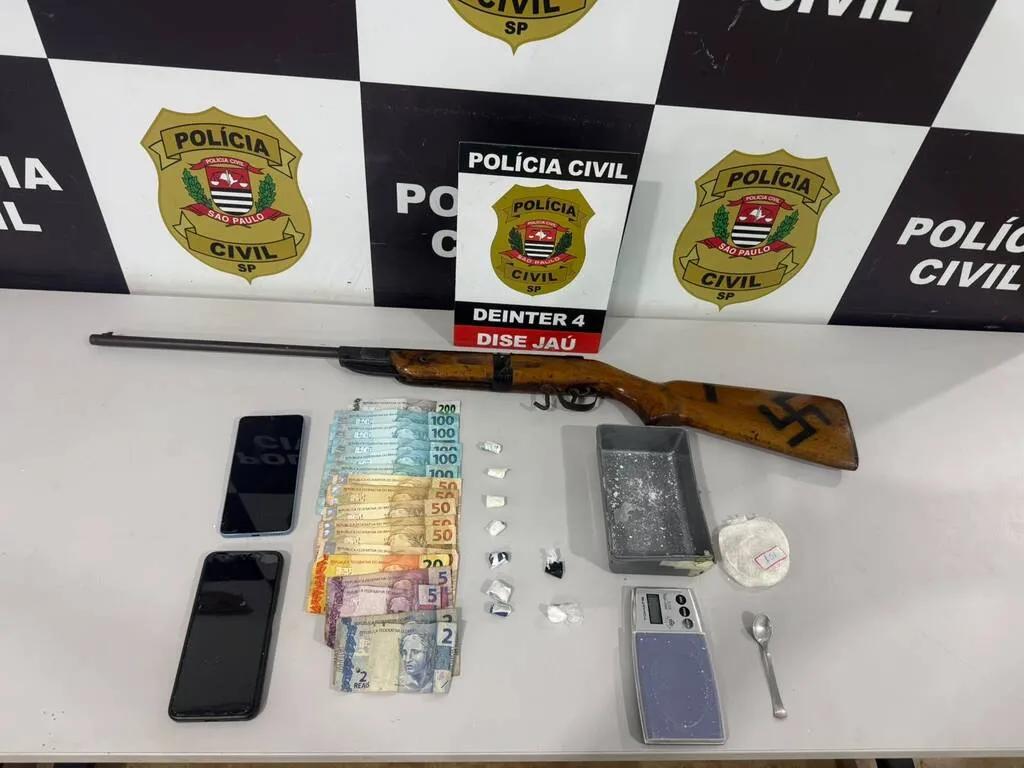 Dinheiro, espingarda de pressão, telefones celulares e balança em cima de uma mesa com painel da Polícia Civil ao fundo