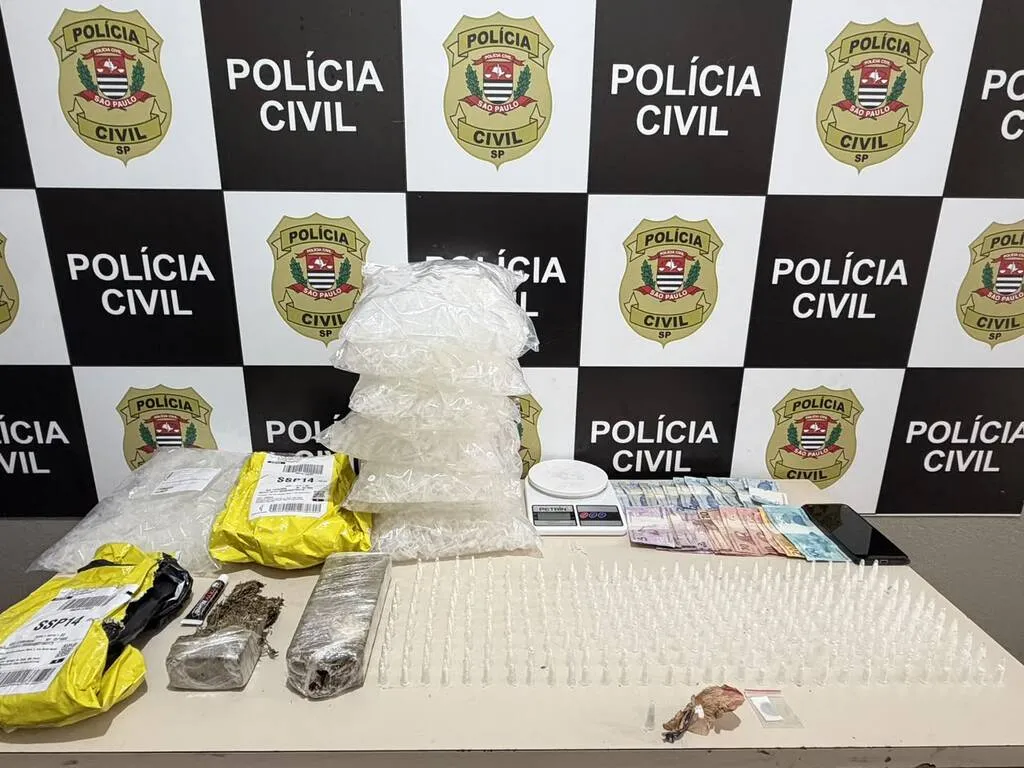 Mesa com dinheiro, embalagens, balança e telefone celular, contendo painel da Polícia Civil ao fundo