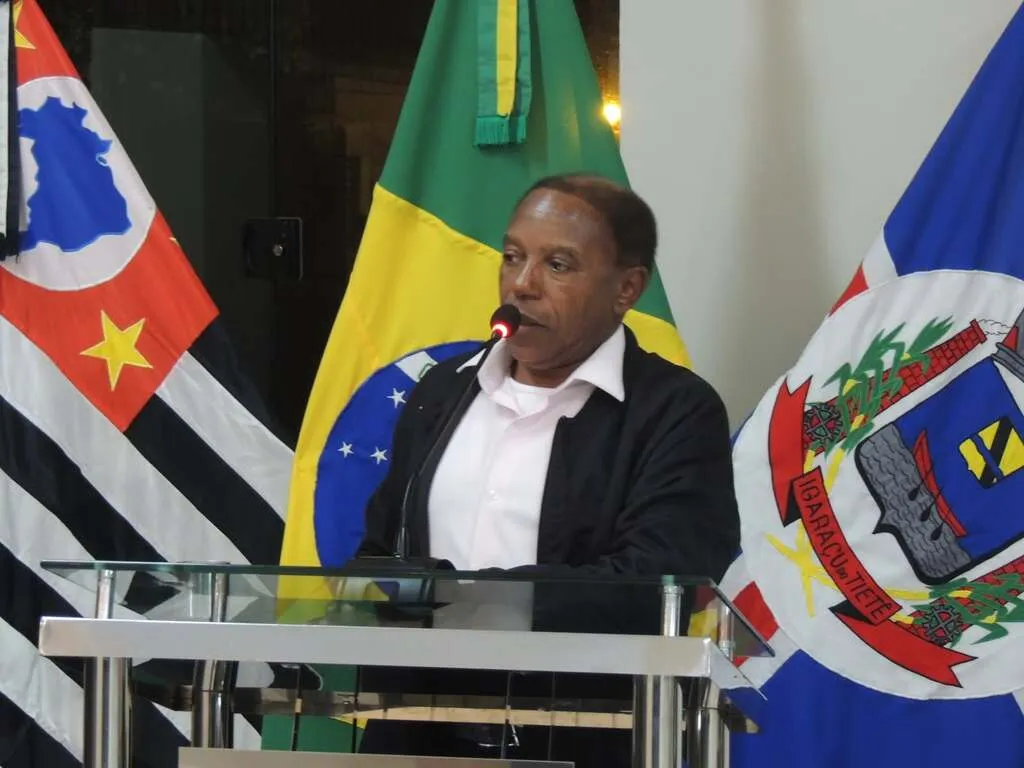 Homem negro discursa em tribuna de vidro, à frente das bandeiras do Brasil, estado de São Paulo e Igaraçu do Tietê